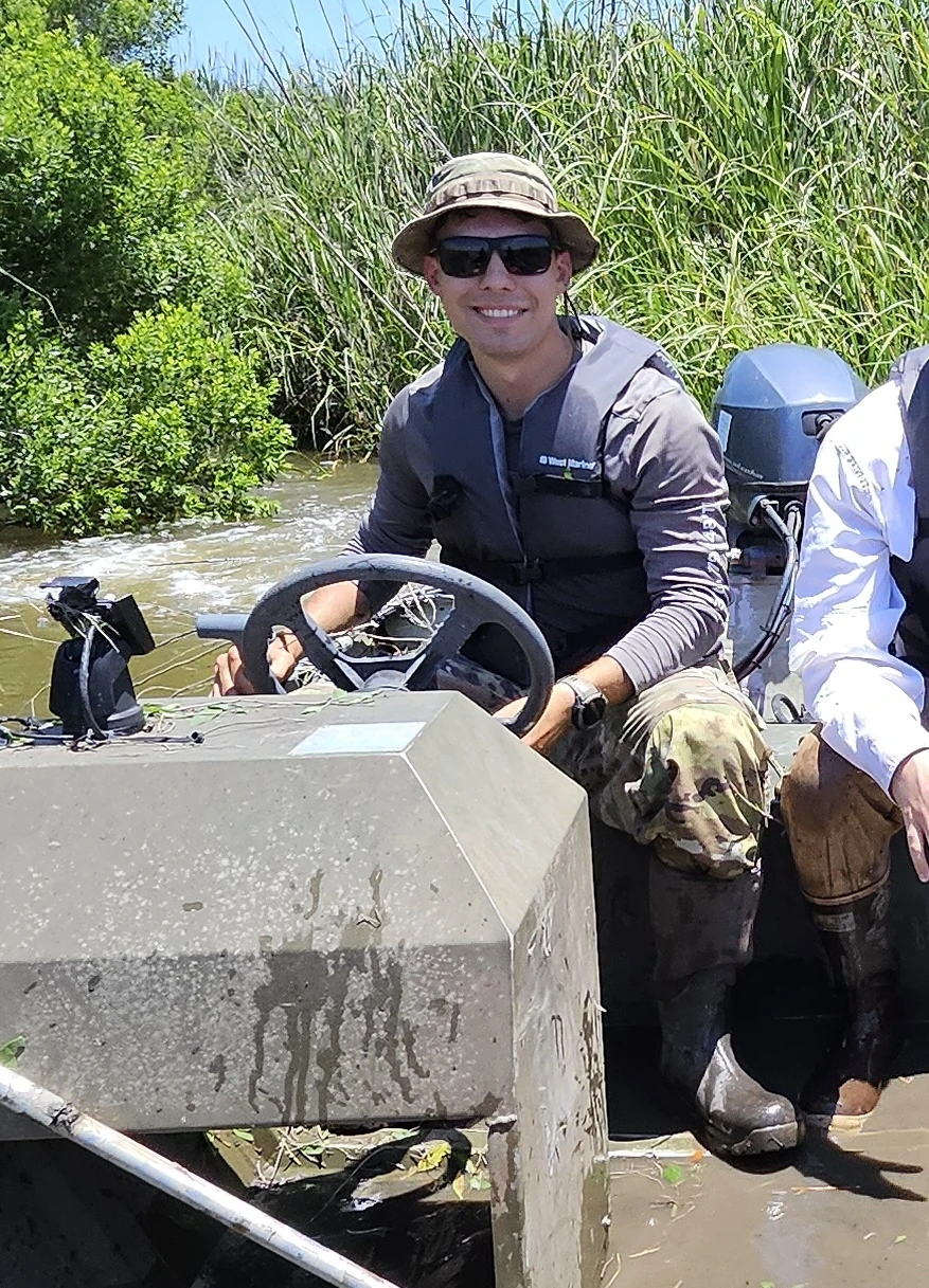 Caleb Yon at the wheel of a boat beside tall marsh vegetation; part of another passenger remains visible at the right edge.